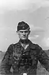 A black-and-white photograph of a man wearing a camouflage military uniform, side cap and a pair of binoculars around his neck. His cap has an emblem in shape of a human skull and crossed bones.