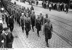 A black and white photograph of a column of Nazi brownshirts marching through the streets of Berlin.