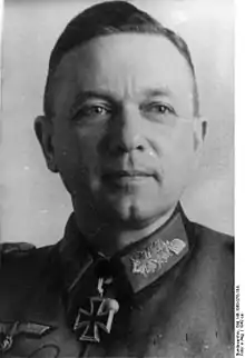 The head of a man, shown in semi-profile. He wears a military uniform with a military decoration in shape of an iron cross displayed at the front of his shirt collar. His hair is dark and combed to back.