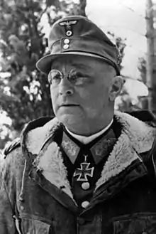 A black-and-white photograph of a man with glasses wearing a military uniform underneath a fur collared coat, field cap and neck order in shape of an Iron Cross.