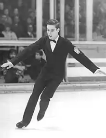 A young male figure skater is performing in an ice rink with a crowd in the background stands. He wears a formal black suit with white shirt and black bow tie, and his short hair is well combed.