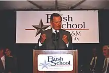 Former president George H.W. Bush speaks at a podium, emblazoned with the Bush School logo, with fists clenched in resolve.