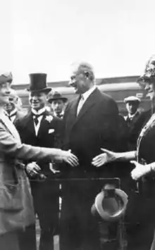 Two white woman extend their hands to one another, as if to shake them, while two middle-aged white men in formal attire look on.