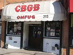 Street view of storefront with doors in the center flanked by windows on both sides.  A white awning says "CBGB" in large red letters in a decorative font, and "OMFUG" below that in the same style.