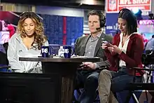 Three people wearing headsets and seated around a table, facing a camera, at a convention