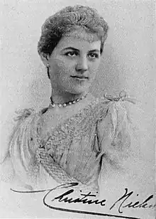 B&W portrait photo of a woman in a light-colored blouse and a choker necklace, with her hair in an up-do; her signature is along the bottom of the photo.