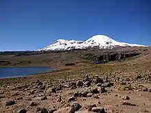 A barren, rock-strewn terrain with two ice-covered mountains in the background; to the left lies a blue lake and to the right a scarp.