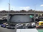 Picture of a road bridge and an avenue with multiple vehicles. Below the bridge lies a giant cone-shaped air vent