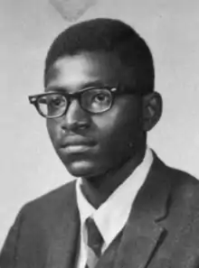 A black and white photo of a boy in a suit, tie, and thick-rimmed glasses, at bust length, staring to the left of the camera