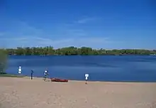 A beach along Cedar Lake with a red and white water safety boat on its sand. Lush green trees line the opposite shore across the deep blue water. A danger sign sticks out of the sand warning people to stay off the lake when it is frozen.