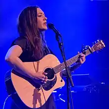 Celine Cairo standing onstage with a blue background, playing an acoustic guitar and singing into a microphone