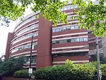 A round, six-story brick building with rows of windows. At its base, there are green bushes and a power line. In the foreground above, there are branches of a tree with green leaves.