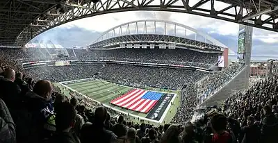 View of Lumen Field during a Seattle Seahawks football game from the stands, overlooking a large American flag covering half of the field, on a cloudy day