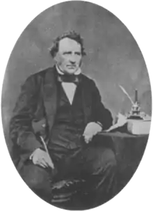 A middle aged Victorian gentleman sits beside a table wearing a dark suit with waistcoat and bow tie. His left elbow rests on the table. In his right hand he holds a quill pen.