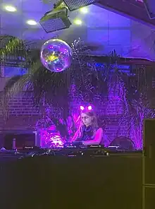  Chelsea Manning, wearing glowing cat ears, workign behind a DJ booth with a discoball overhead