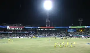 View of a cricket stadium. It has some players in yellow outfit and they are folding yellow flags.