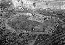 Large circular depression outlined by a stone wall. The bottom is flat and grassy, and has a collection of rectangular stone foundations and smaller circles of stone. A great sandstone cliff towers in the background, and beneath the cliff are other stone foundations that are larger and higher.