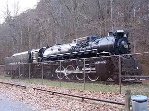 A locomotive with chain link fencing in the foreground and a forested ridge in the background.