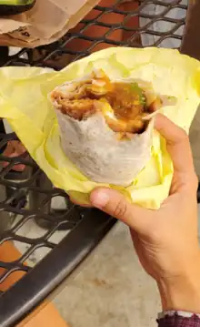 A photograph of a hand holding a chile relleno burrito wrapped in yellow paper in front of a brown metal picnic table.