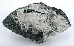 Photograph of a mendipite sample with a dark crystal of chloroxiphite embedded in it and a smaller, bright blue diaboleite crystal at the chloroxiphite's top edge.