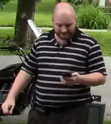 Still image from a video of Cantwell, who is wearing a black and white striped polo shirt and looking down at a cell phone in his hand.