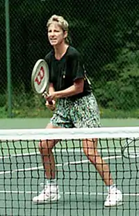 A blonde-haired female tennis player with multi-coloured shorts and a black shirt, with the tennis racket out in front of her