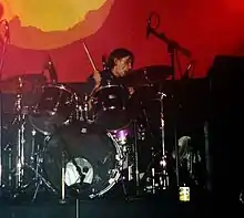 A man performing on a purple drum kit against a red-and-yellow backdrop. A drum stick is visible in his right hand; several microphones and a cooling fan are also visible.