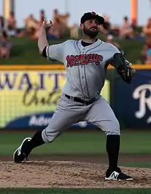 A man in a gray baseball uniform with "Nashville" written on the chest in red, high black socks, and a black cap holds a ball in his right hand high behind his head in mid-pitching motion.