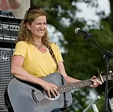 A woman wearing a yellow t-shirt smiling and playing a black acoustic guitar.