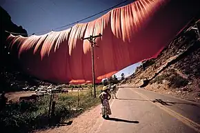 An enormous volume of fabric hangs from a wire across a valley. In the foreground is a telephone pole and several people looking up.