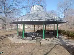 An open-air gazebo.  The structure is eight-sided, with a metal roof supported by pillars with decorative iron fencing between the pillars.  Under the roof are permanently installed tables with attached seating for about a dozen people.