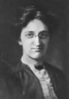 A young white woman with dark hair and eyes, wearing eyeglasses and a dark dress with a light ruffled front section