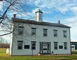 A two-story five-bay clapboard-sided building in white with green trim, topped by a gabled roof with two small brick end chimneys and a tall cupola in the center