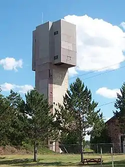 A large grey building, wider at the top that towers over the pine trees in the foreground