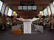 The interior of a building with a black sign suspended from the ceiling reading "PLEASE WAIT IN THIS AREA UNTIL PLATFORM IS INDICATED"