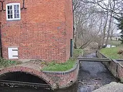 Close view of the south side of the building.  A small eliptical brick arch leads the water into the enclosed millrace, and there is an overflow weir to the right of the building. Trees are visible on the riverbank beyond.  The water is constrained in a red brick channel with a bulnose (curved) corner.  Two scroll shaped iron 'plates' are on the upper wall, terminals for tie-rods that pass through the building. This is a winter view, and the state of the trees makes this stark. There is a small and unturbulent flow of water through the overflow weir, because the level is accurately controlled by the main weir in the old lock chamber. A precarious plank bridge, with no handrails, crosses the overflow race to reach the lockside on the right.