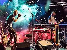 A man sings with a microphone on the stage while another one plays the bass, the background has blue and red lights, while confetti is being released as well