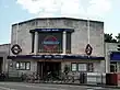 A light-grey-bricked building with a blue sign reading "COLLIERS WOOD STATION" in white letters all under a blue sky with white clouds