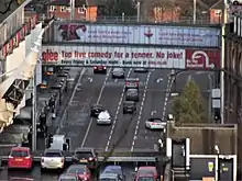 High level shot of multi lane road lower down stretching into distance with two glass sided pedestrian walkways over linking car park to right and shopping centre to left