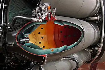 Rolls-Royce Nene with nine combustion chambers. The cutaway is one of 2 chambers fitted with a flame igniter which places the igniter in a cooler location than directly in the hot gas stream. During a start atomized fuel from the small self-contained unit (orange-coloured solenoid shown) is ignited by its ignition plug and the flaming jet of fuel is projected into the main fuel spray from the burner. Combustion is propagated to all the chambers through interconnecting tubes.