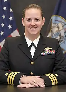 A white woman in a black US Navy uniform is sitting at a table with her hands folder upon it; she is smiling and looking into the camera.