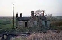 A photograph of the now demolished railway station at Constable Burton in Wensleydale, Yorkshire, England.  A single railway line runs in front of the building. Two boarded-up windows are shown, as well as an open door which led into the old booking office. Four chimney stacks, some with large ornate Victorian chimneypots are on the roof. A telegraph pole stands to the left of the station.