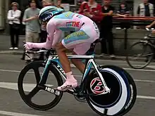 A man in pink clothes and pink shoes, sitting ducked down on a bicycle. In the background people are watching.