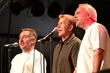 Three men singing on stage into microphones.