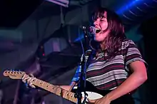 A female singer and guitarist performing onstage. She is singing into a microphone while playing electric guitar.