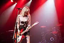 Woman onstage, holding guitar and looking down, smiling