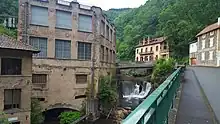 Recent shot of the former Creux de l'enfer factory taken from the opposite bank. On the right is the avenue, in the center the Durolle with its waterfall under a modern bridge, on the left the 3-storey factory facade. Part of the factory is built on vaults above the water, the plaster on the walls is crumbling, revealing the shale stones in places, and the name, painted in red, is still visible