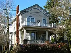 Photograph of the Cross House, a two-story house with a broad, two-level porch, bay windows on the front and side, and a brick chimney, seen from the front from a level below the foundation of the house