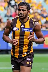 A male athlete with dark hair wearing a sleeveless jersey and shorts runs on the grass surface of the playing arena.