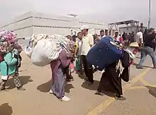 Three women in Moroccan attire carrying bales on their backs.